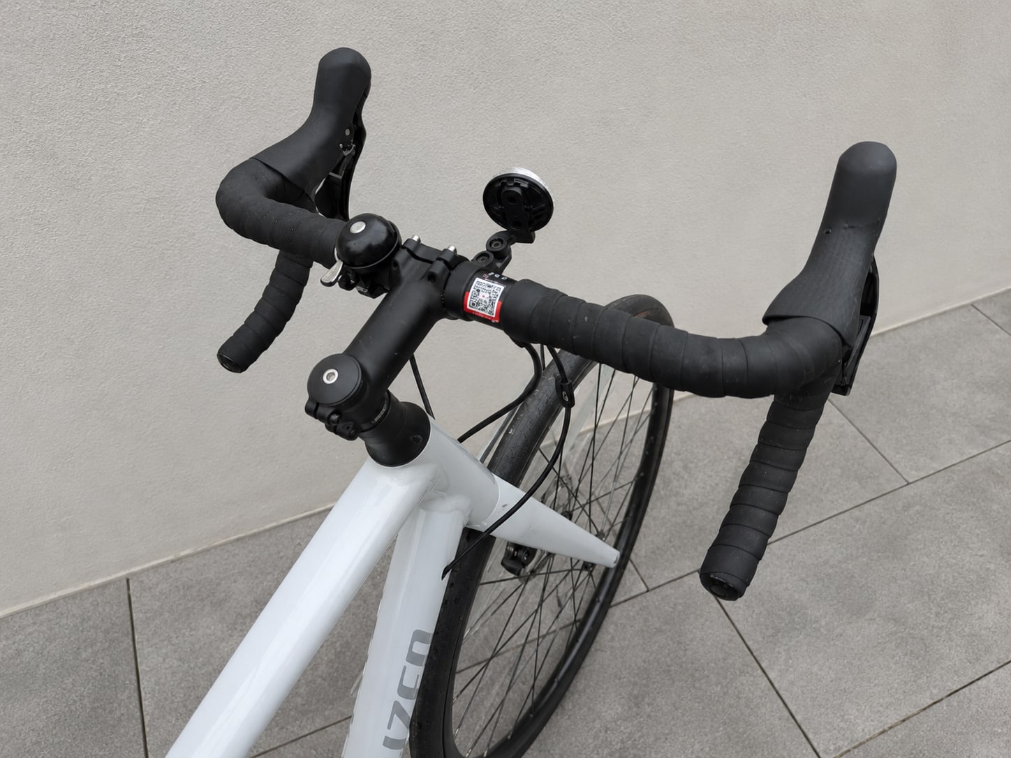 White and black drop-bar road bike, non-drive side, overhead angle against tiled pavement and grey wall.