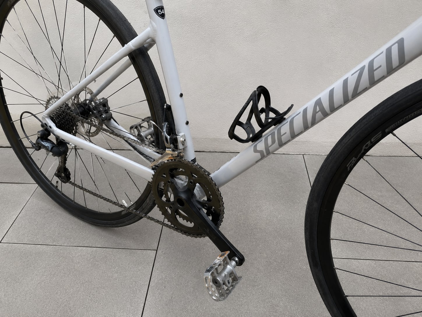 White Specialized road bike drivetrain detail, drive side, on concrete floor. Close-up of crankset, chain, cassette, and rear derailleur.