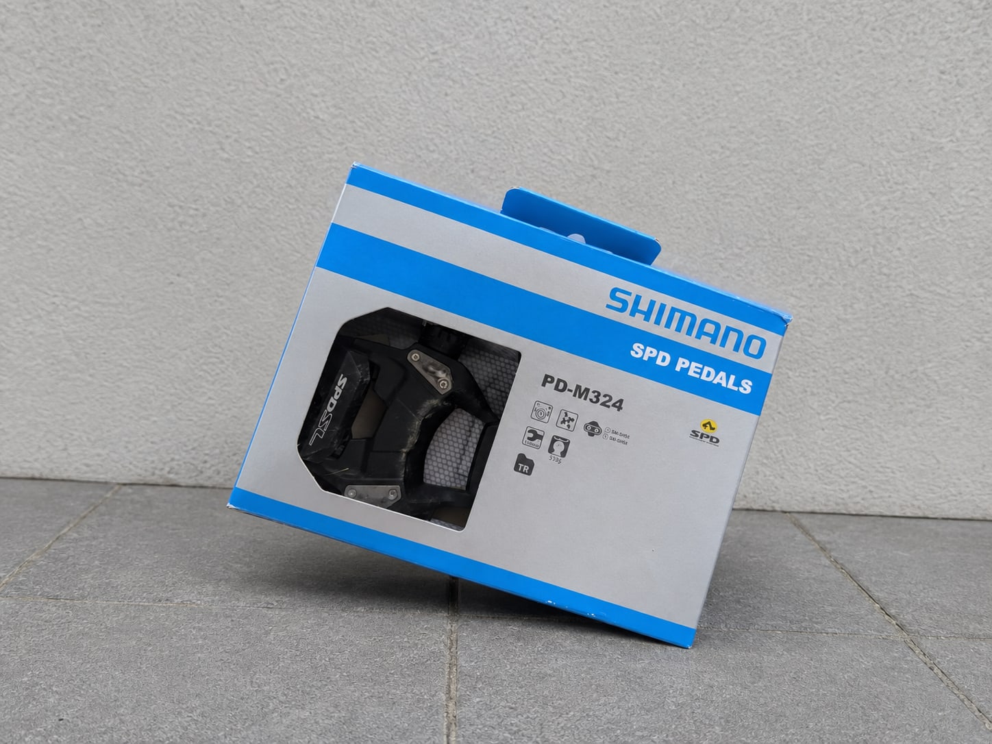 Shimano PD-M324 SPD pedals in retail packaging, black with grey accents, displayed in product box window against grey concrete background.
