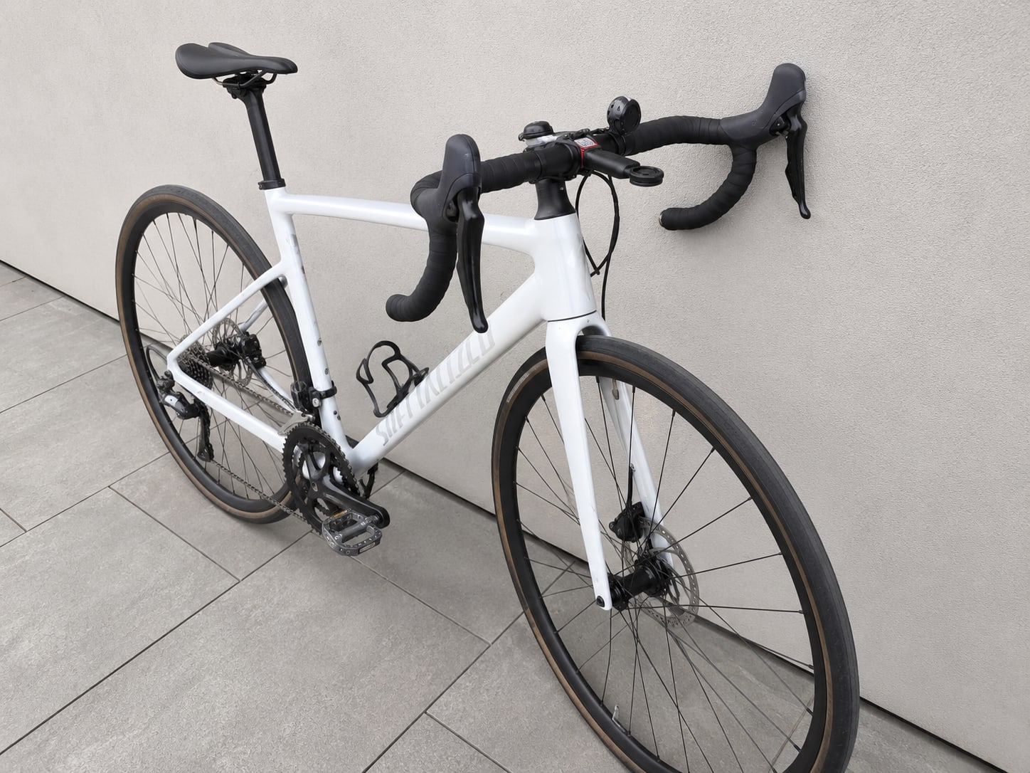 White gravel bike with black drop bars and brown-wall tyres, three-quarter view from drive side, on tiled floor against white wall.