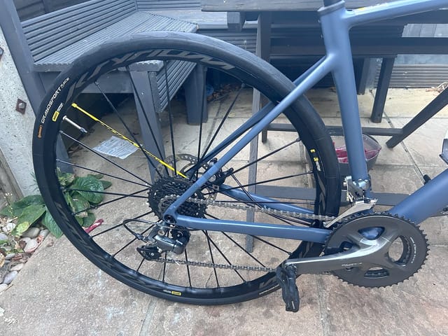 Blue gravel bike on concrete, drive side view, front wheel prominent, frame and drivetrain visible, metal structures in background.