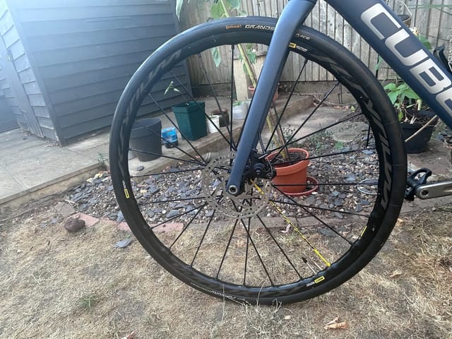 Front wheel of blue gravel bike with Continental Grand Prix tyres, close-up on drive side, outdoor garden setting with shed and gravel ground.