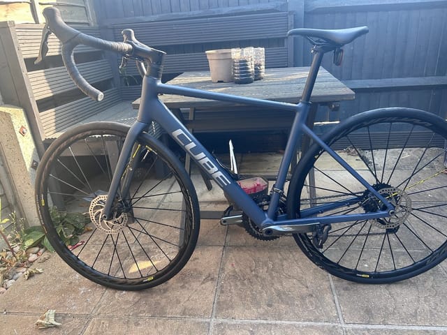 Blue Cube road bike with drop bars, parked on concrete outside a metal shed, drive side view, with red rear light visible.