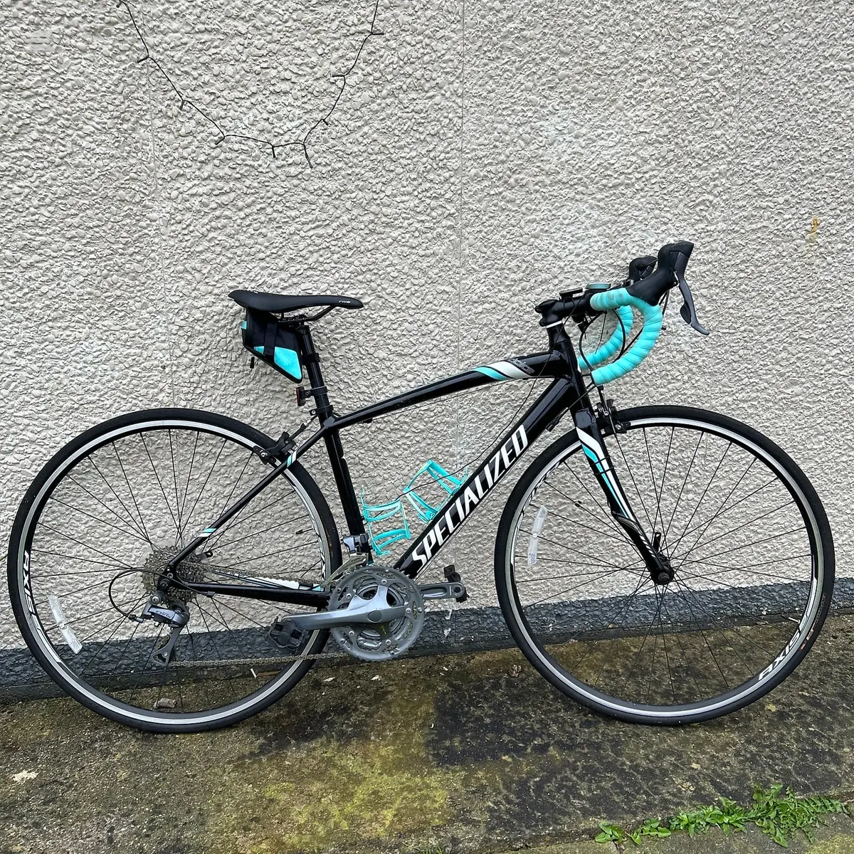 Black and turquoise Specialized road bike against a textured concrete wall, drive side view, with turquoise bar tape and saddle bag.