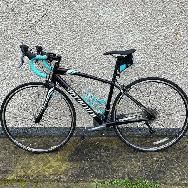 Black and turquoise Specialized road bike against a textured concrete wall, drive side view, leaning against building.