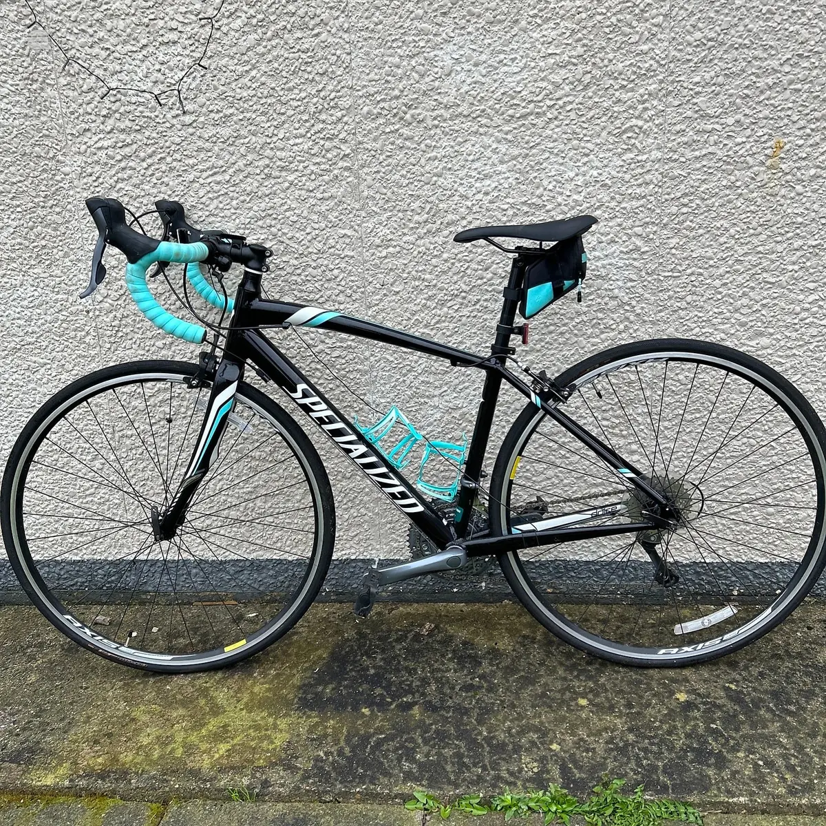 Black and turquoise Specialized road bike against a textured concrete wall, drive side view, leaning against building.
