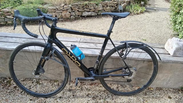 Black Orro road bike with blue water bottle, drive side, resting on wooden bench against gravel driveway and stone wall.