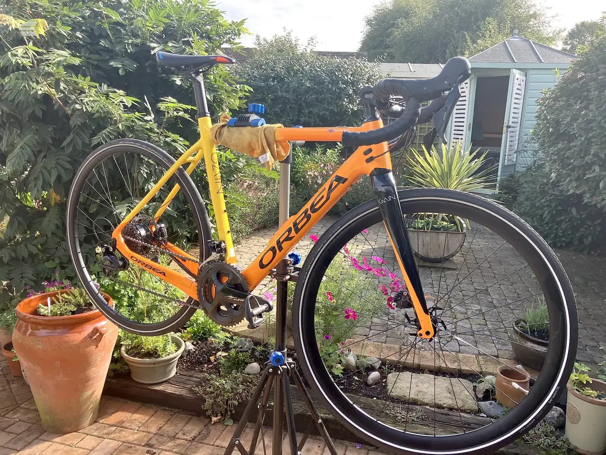 Orange and yellow Orbea gravel bike on a stand, drive side, in a garden patio setting with potted flowers and a shed.