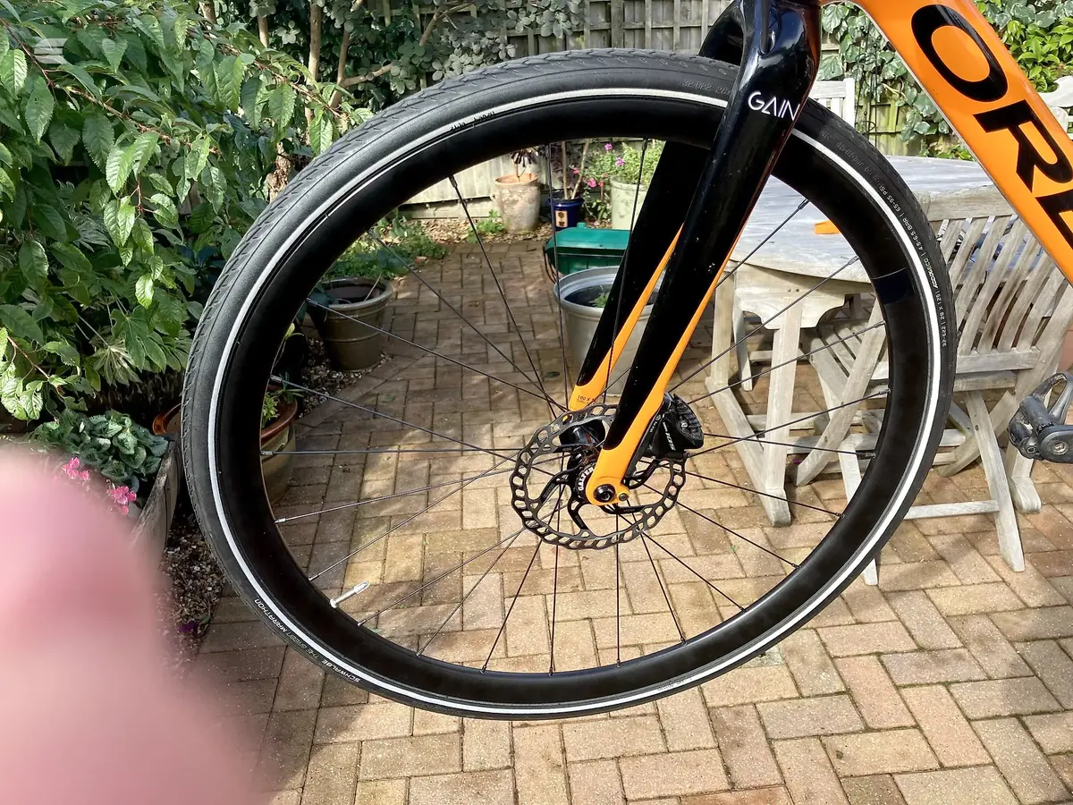 Orange and black gravel bike, front wheel close-up, non-drive side, in a garden patio setting with wooden furniture and potted plants.