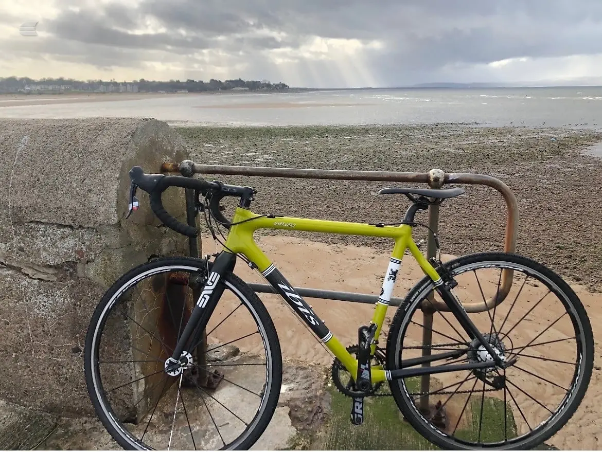 Lime-green and black road bike leaning against a concrete block on a beach, drive side, with sea and cloudy sky behind.
