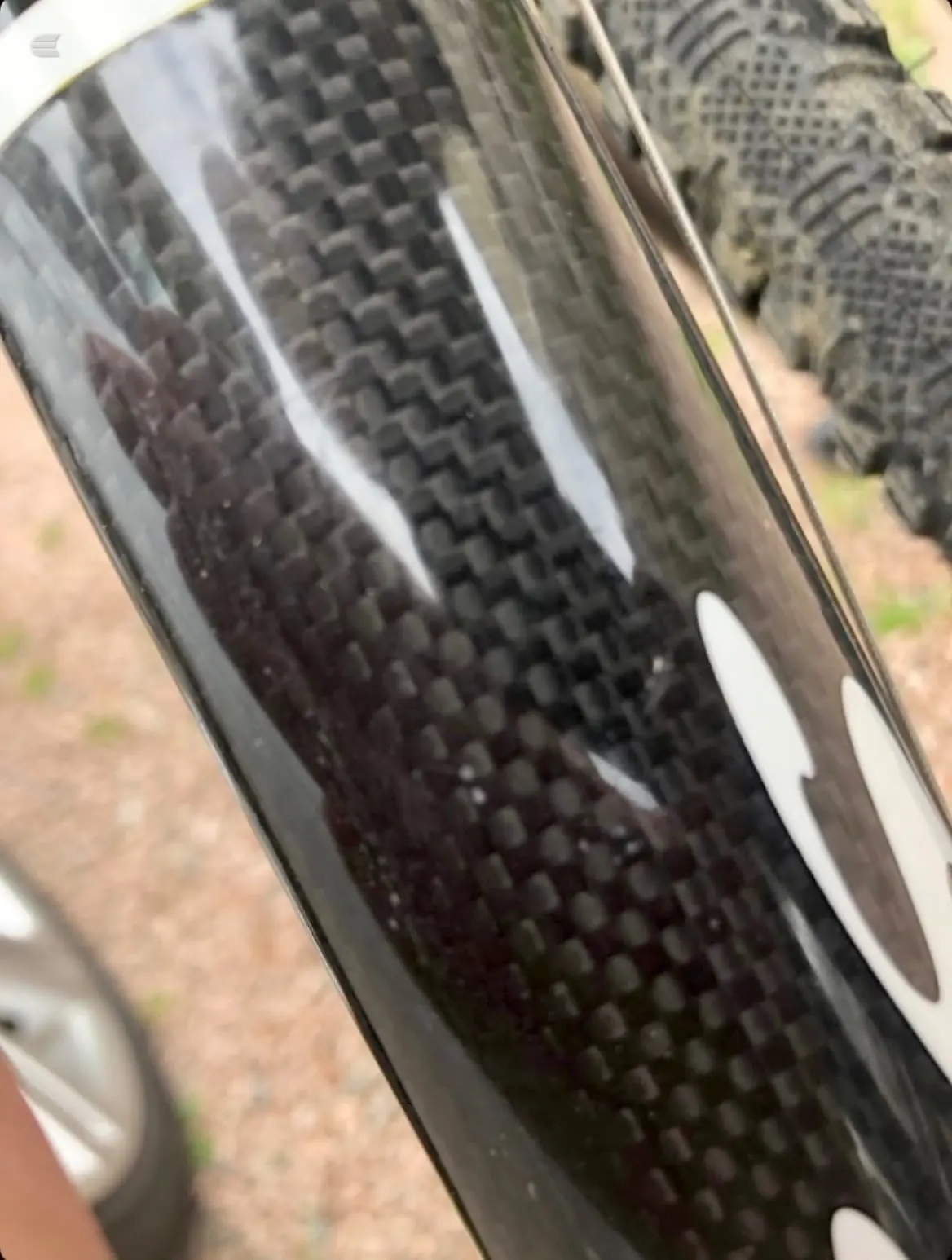 Close-up of a bicycle wheel with black tyre and carbon fibre rim, angled view showing tread pattern and rim detail, with blurred bike frame and ground in