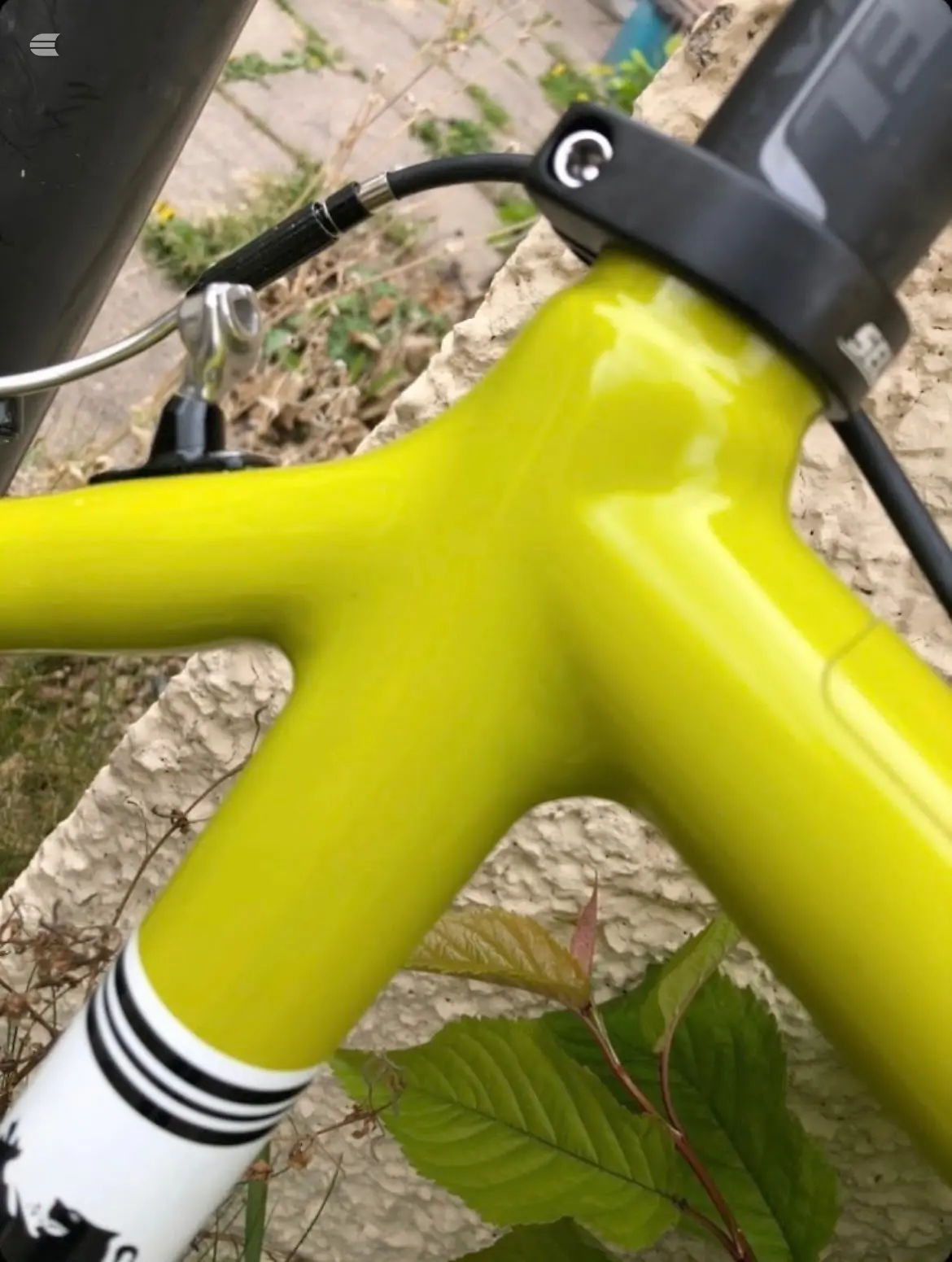 Close-up of bright yellow electric bike frame and headtube with black hydraulic disc brake hose and stem connector, on concrete ground with weeds.