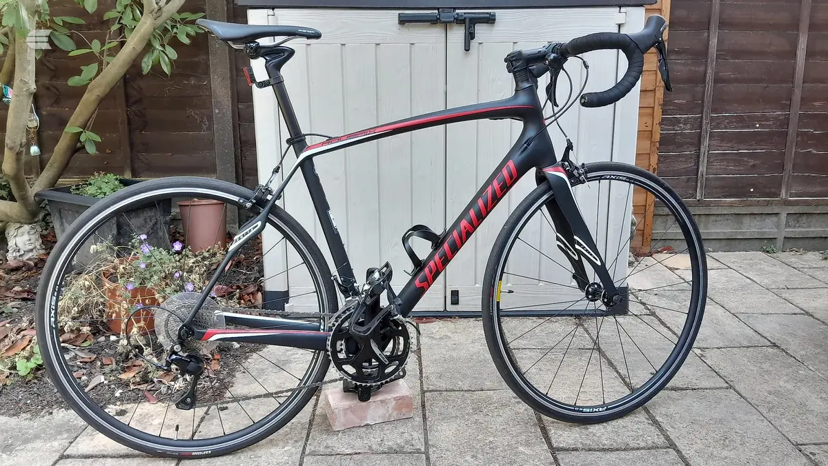 Black and red Specialized road bike on paved driveway, drive side, against wooden garage doors with potted plants nearby.