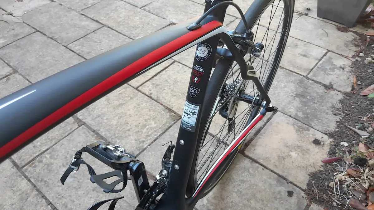 Black and red road bike on paved ground, drive side view, with drop bars and cleats visible.