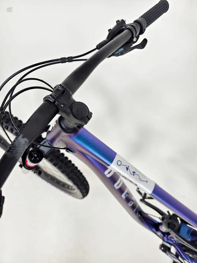 Purple and white hardtail mountain bike, front-left 3/4 view, handlebars and front wheel prominent, against plain white background.