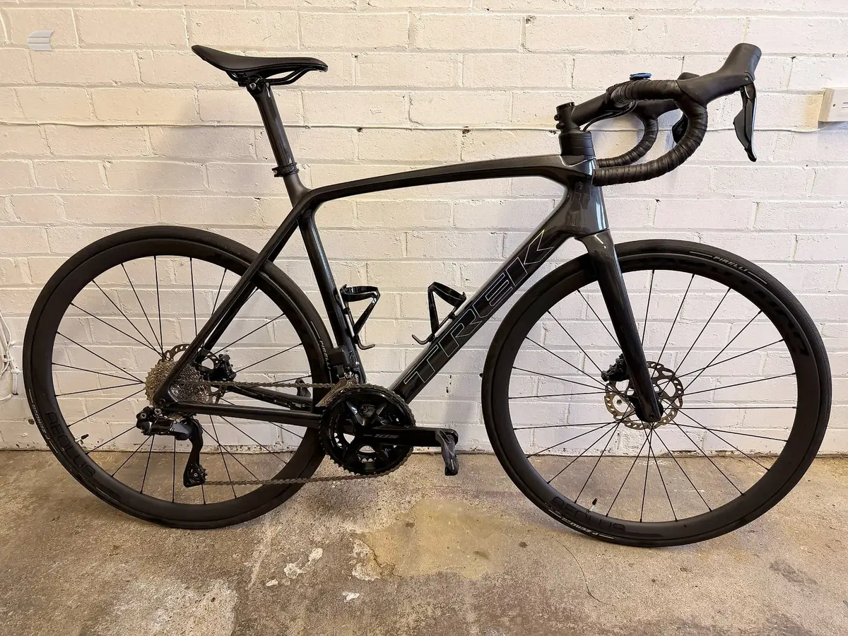 Black Trek road bike with disc brakes, drive side, leaning against a cream brick wall on concrete ground.