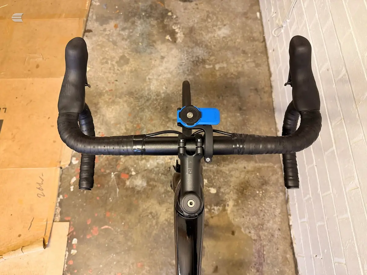 Black drop-bar bike viewed from above on concrete and wooden flooring, showing handlebars, stem with blue computer mount, and frame.