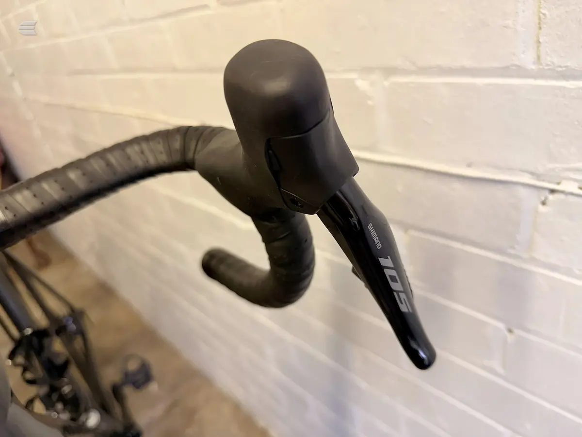 Black Shimano drop bar brake lever and wrapped handlebar on a road bike, close-up against a white brick wall.