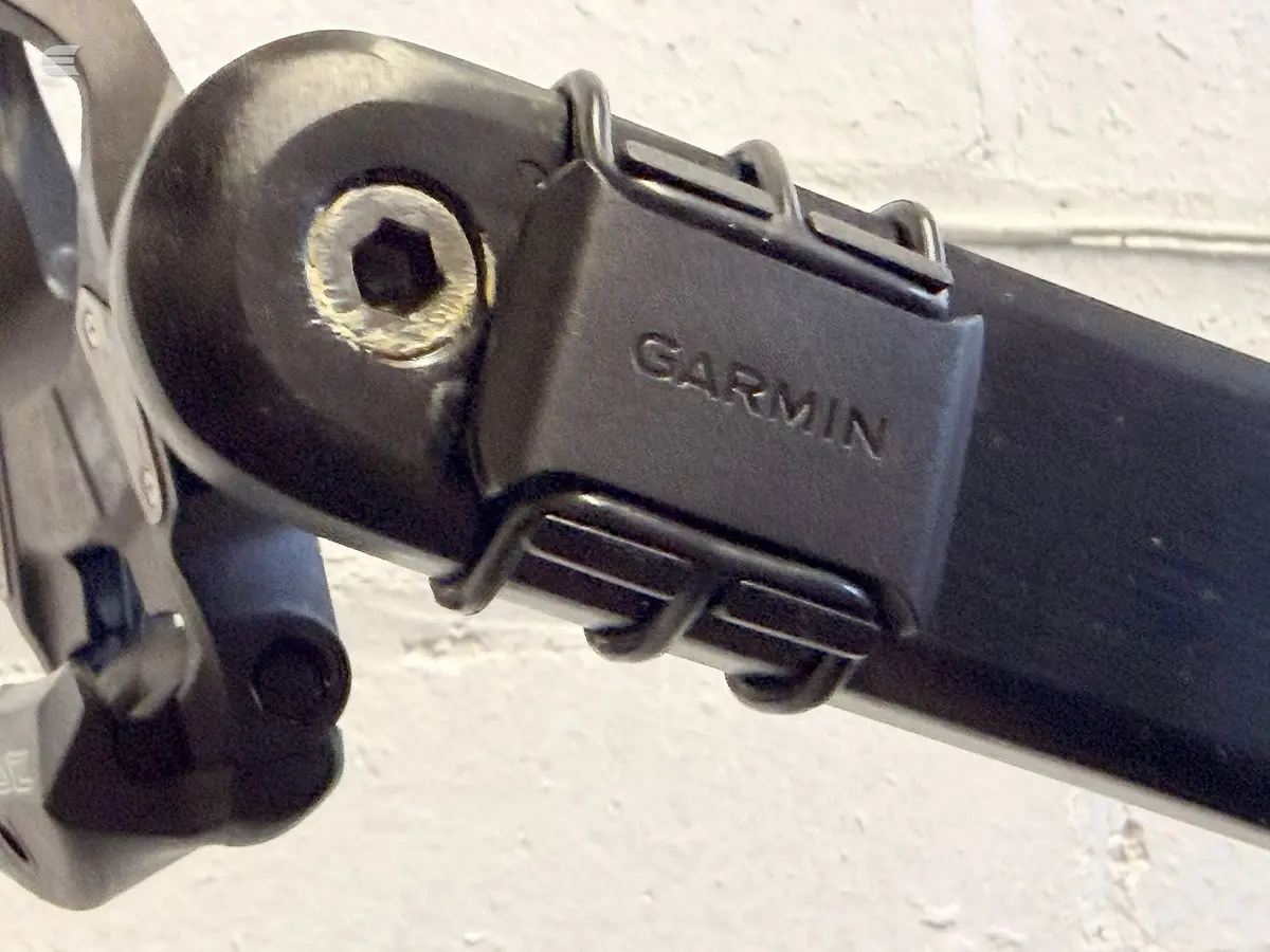 Close-up of Garmin bike computer mounted on handlebar stem, black plastic housing with hexagonal attachment points, white concrete wall background.