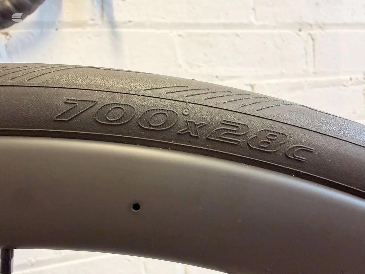 Close-up of a Zoox 28C bicycle tyre on the rim, showing embossed branding and tread pattern against a white brick wall.