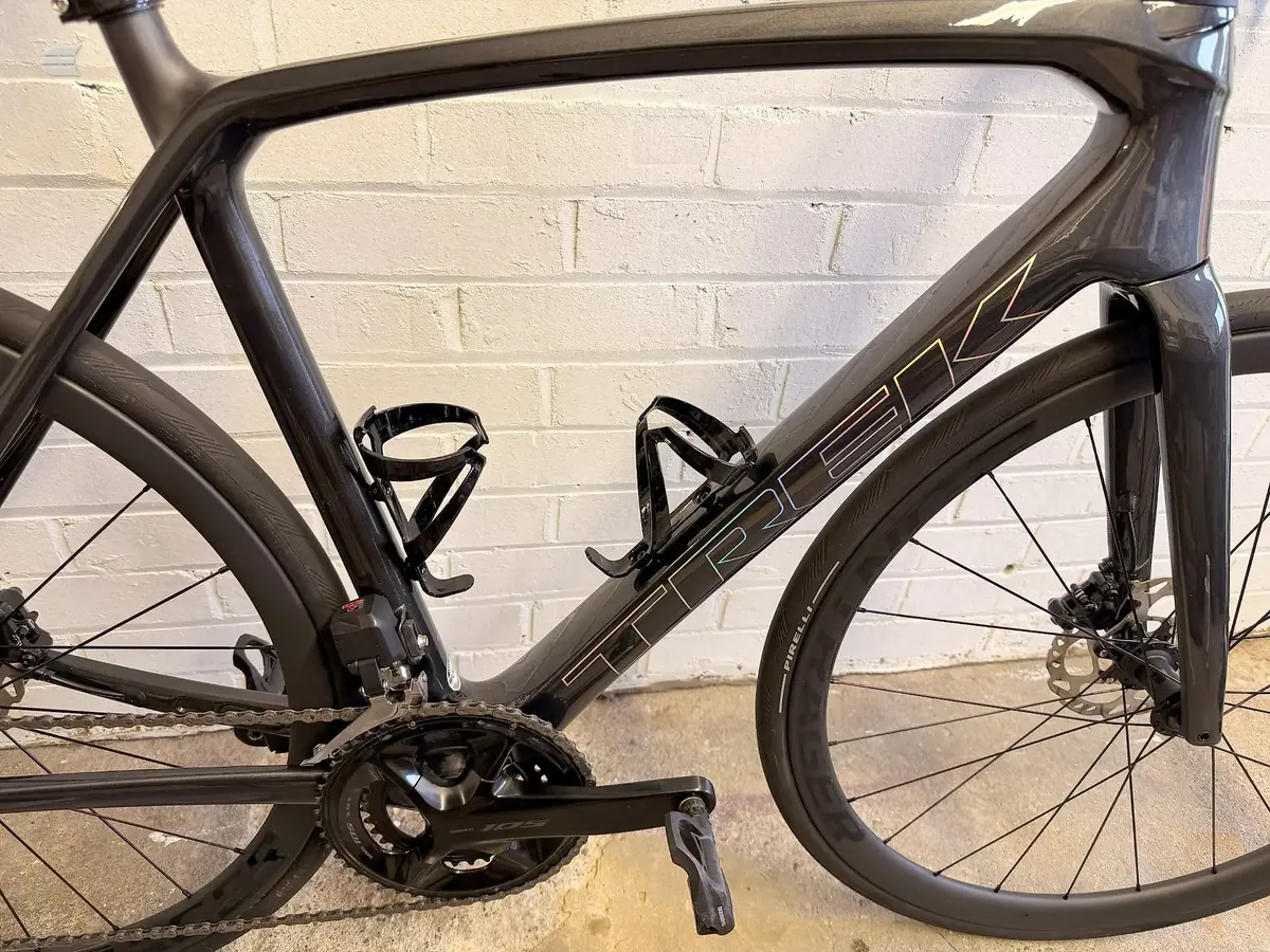 Black road bike on a stand, drive side, against a cream brick wall. Frame shows gold accents on the down tube.