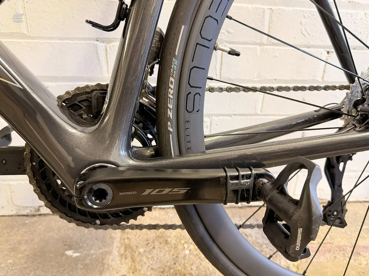 Close-up of Shimano 105 crankset and chain on a grey road bike, drive side, against a brick wall.