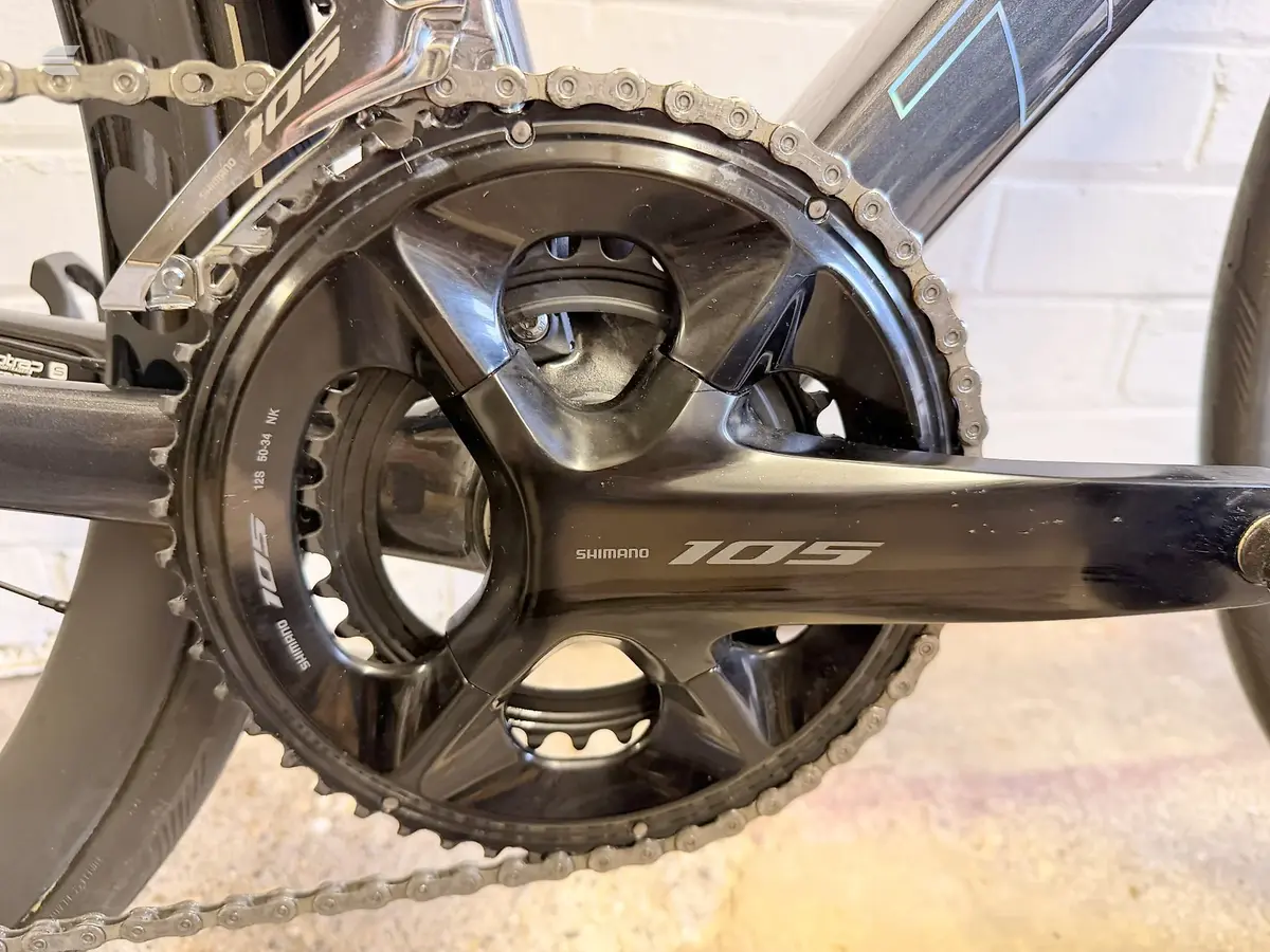 Shimano 105 crankset and chainrings on a black road bike, drive side close-up against a brick wall.