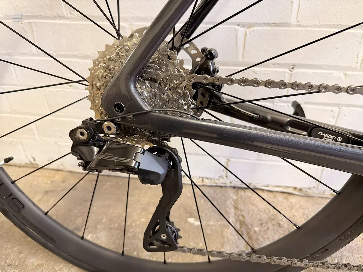 Shimano rear derailleur and cassette on black road bike, drive side close-up against brick wall.