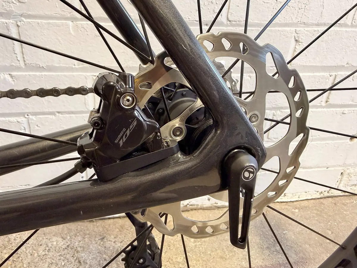 Shimano hydraulic disc brake and rotor on black mountain bike, drive side close-up against white brick wall.