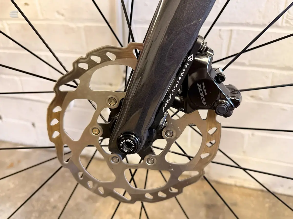 Shimano hydraulic disc brake and rotor on bicycle fork, close-up detail shot against white brick wall background.
