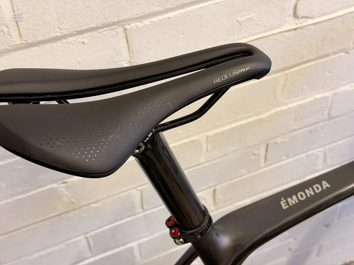 Black Heullez Comp saddle on Émonda bike frame, seatpost detail, against white brick wall.