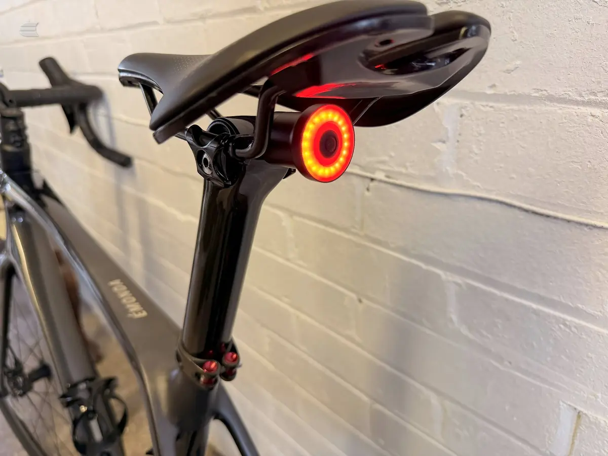 Close-up of a black bicycle seatpost and saddle with a bright red circular rear light mounted below, against a white brick wall.