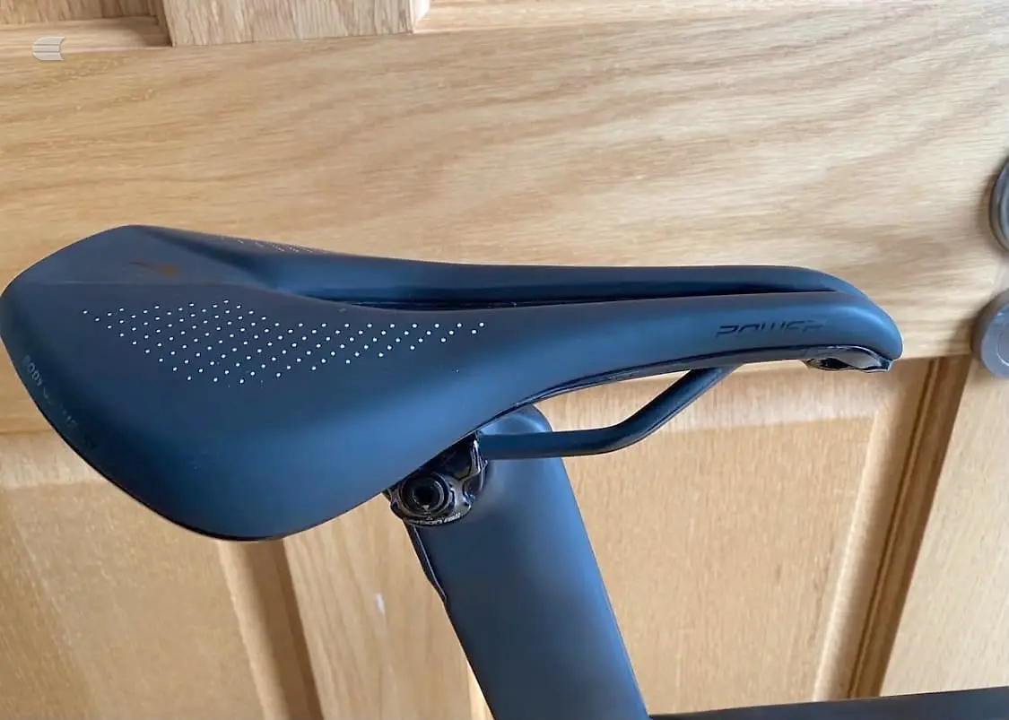 Black saddle with perforated top panel on seatpost, wooden floor background, non-drive side view.