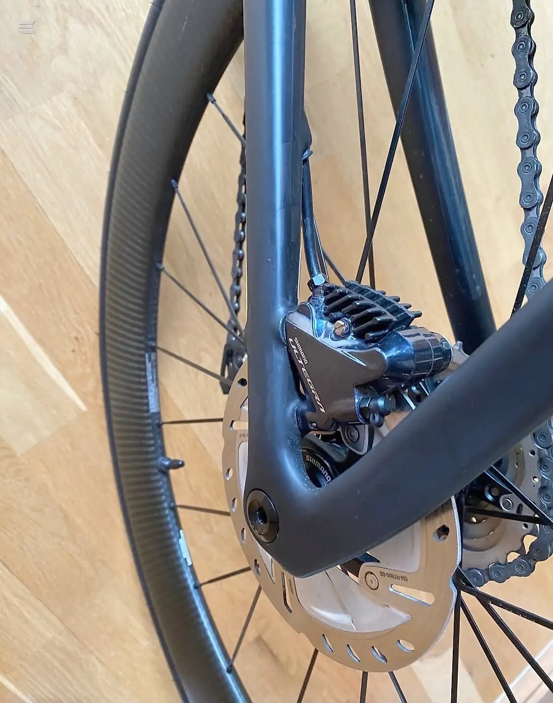 Close-up of disc brake caliper and chain on blue-frame bike, drive side, indoors on wooden floor.