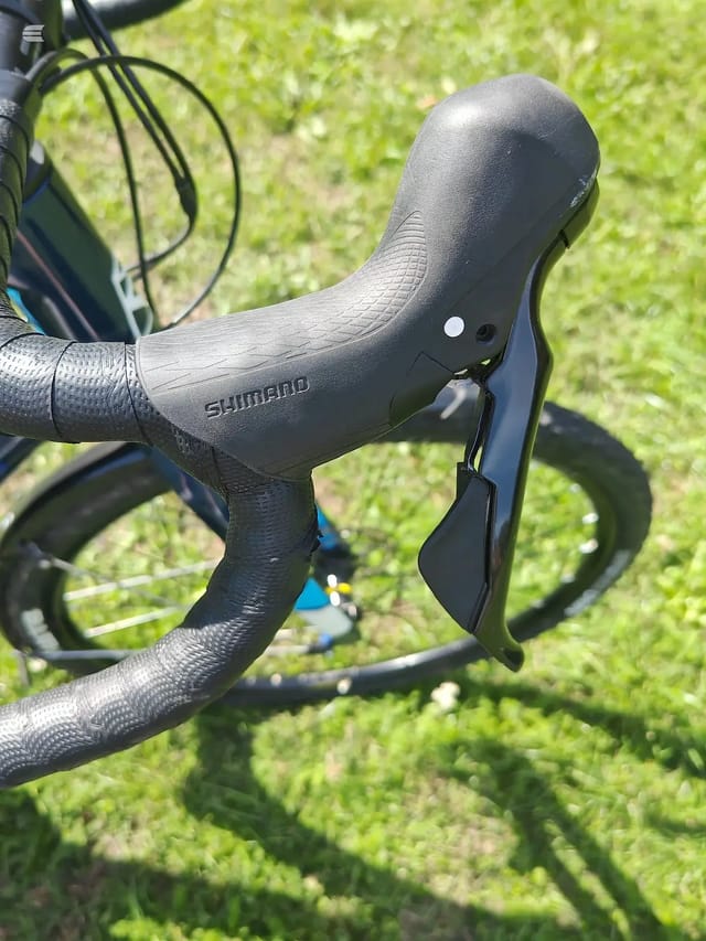 Shimano drop bar brake lever and shifter on black drop bars with textured grip tape, mounted on a road bike in bright sunlight over green grass.