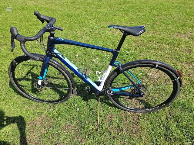 Blue and white gravel bike on grass, drive side, three-quarter view. Drop bars, disc brakes, mudguards fitted.