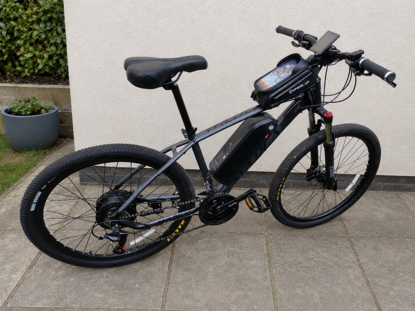Black hardtail e-bike with fat tyres, front suspension, and integrated smartphone mount on handlebars, parked on paved ground against a white wall, drive side