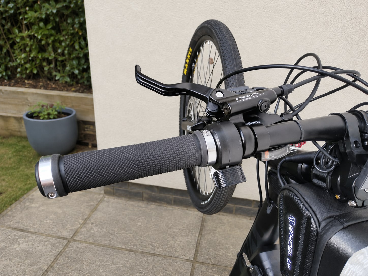 Close-up of black mountain bike handlebars and brake levers, non-drive side, with front wheel visible behind, on a paved patio with garden in background.