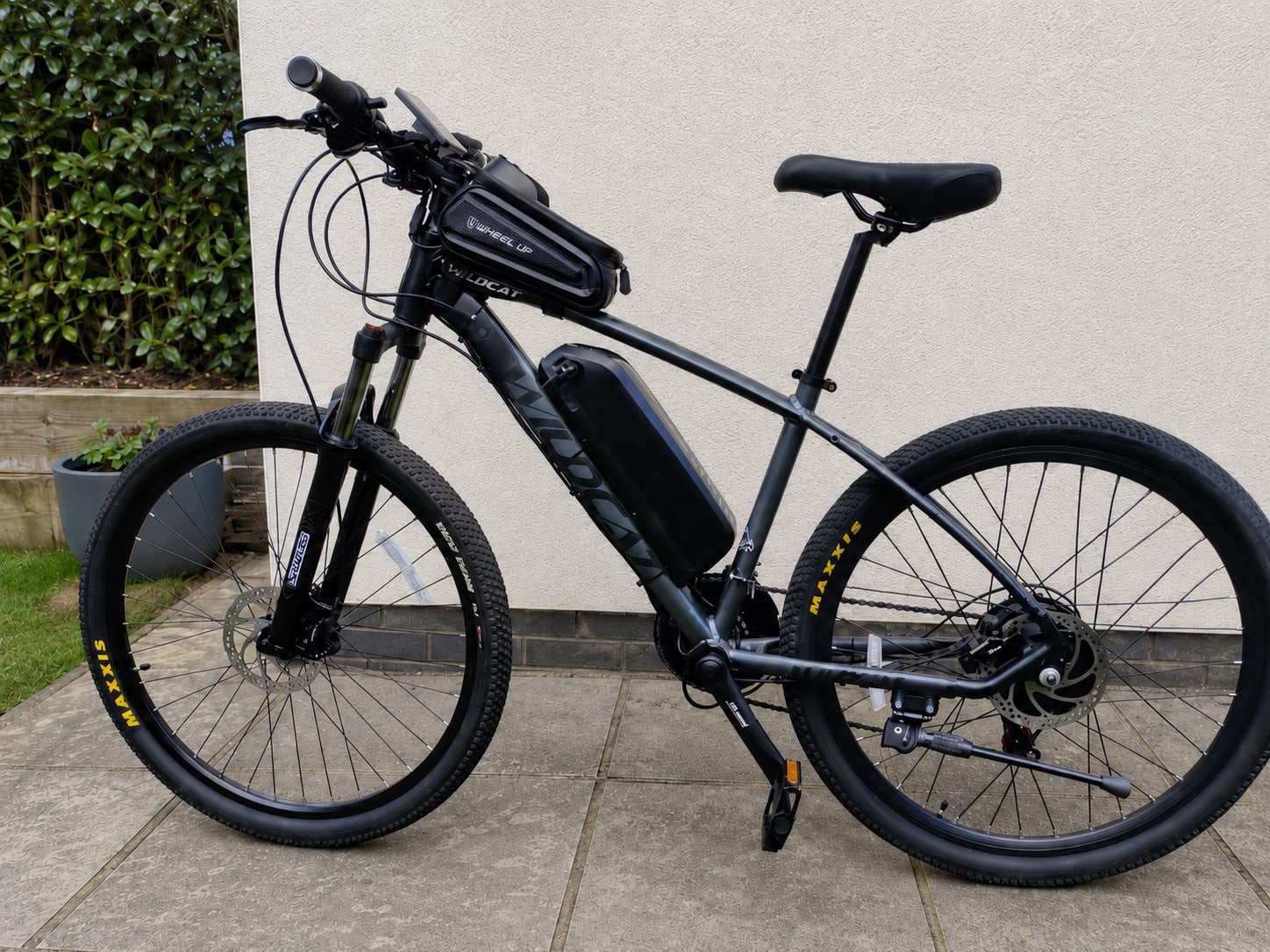 Black hardtail e-bike with blue accents, drive side, on paved ground against a white wall with ivy and grass visible.