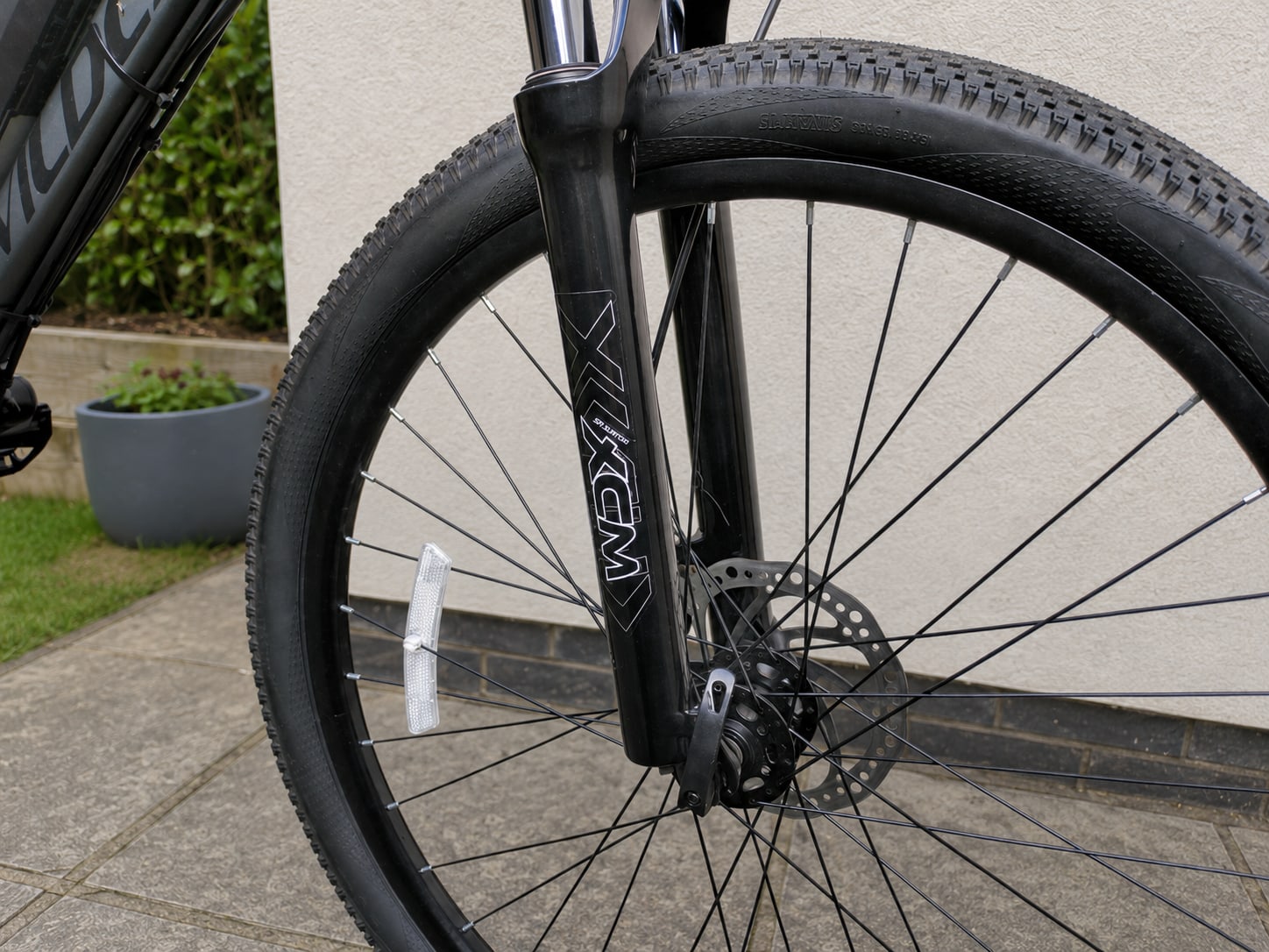 Black gravel bike front wheel with disc brake, close-up on non-drive side, parked on paved driveway with potted plant and hedge in background.