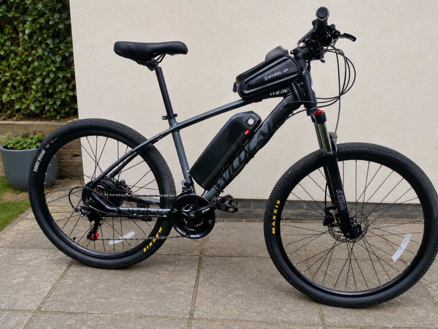 Black electric mountain bike with full suspension, drive side, on paved driveway against white wall with ivy planter.