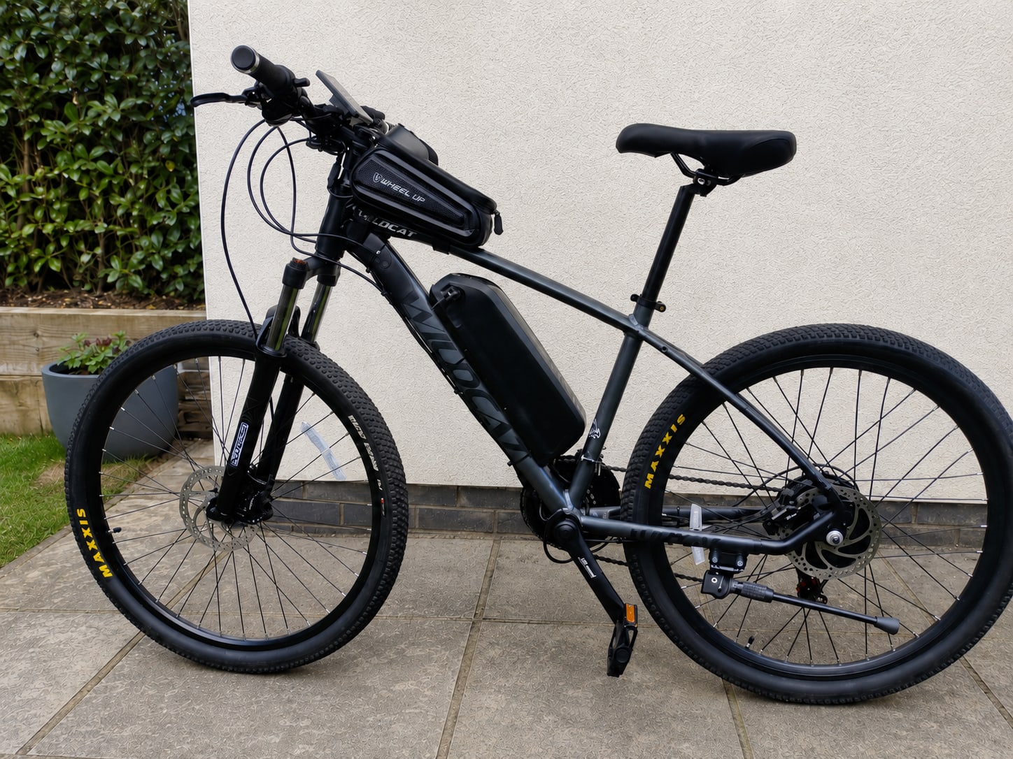 Black electric mountain bike on paved driveway, drive side, against white wall with ivy and planter.
