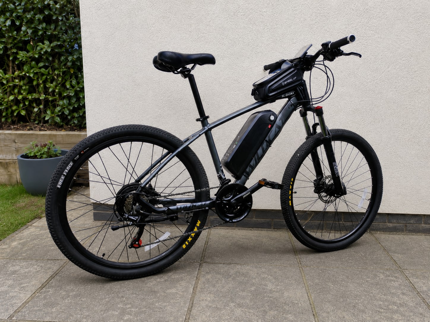 Black and grey electric mountain bike on paved driveway, drive side, against white rendered wall with ivy and potted plant.