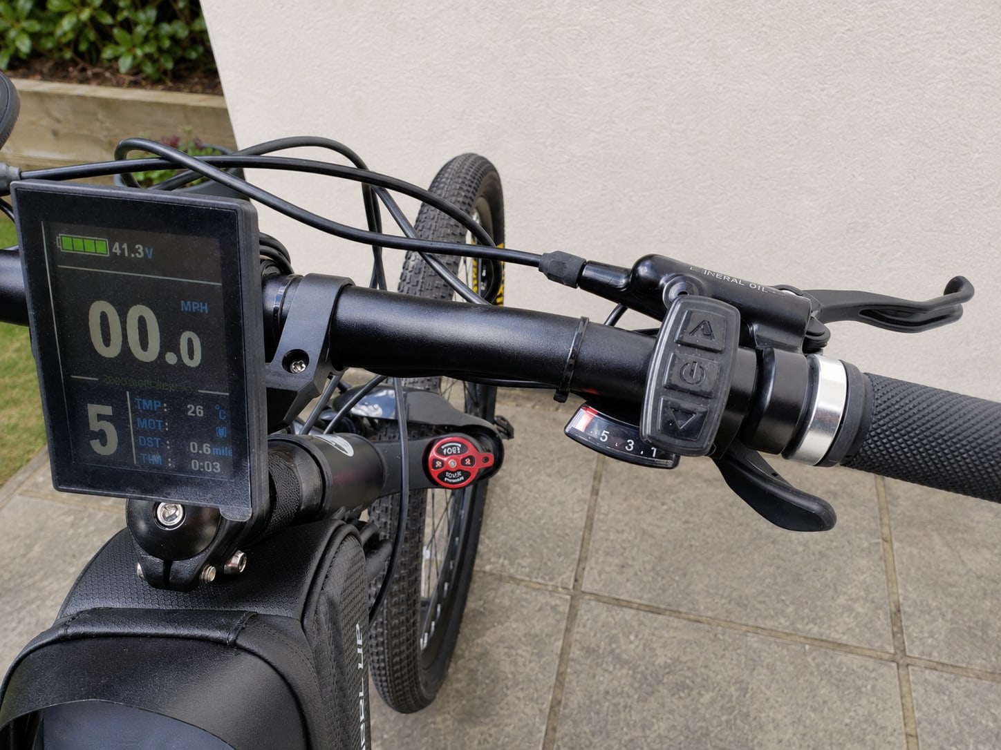 Electric bike cockpit detail showing digital display with battery indicator and speed readout, handlebars with brake lever and throttle control, against a