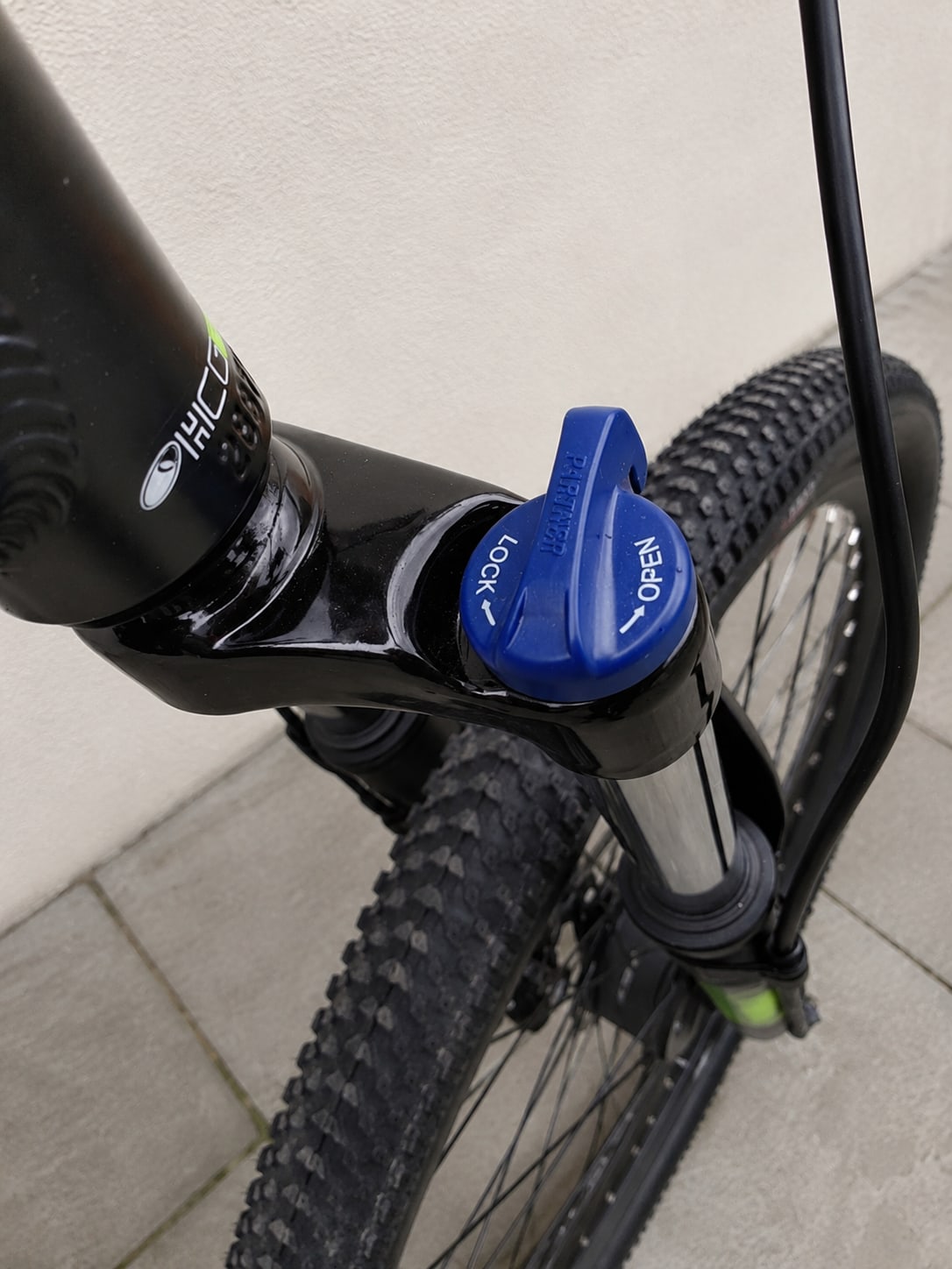 Close-up of blue brake lever marked 'OPEN' and 'LOCK' on black mountain bike handlebars, non-drive side, indoors against white wall.