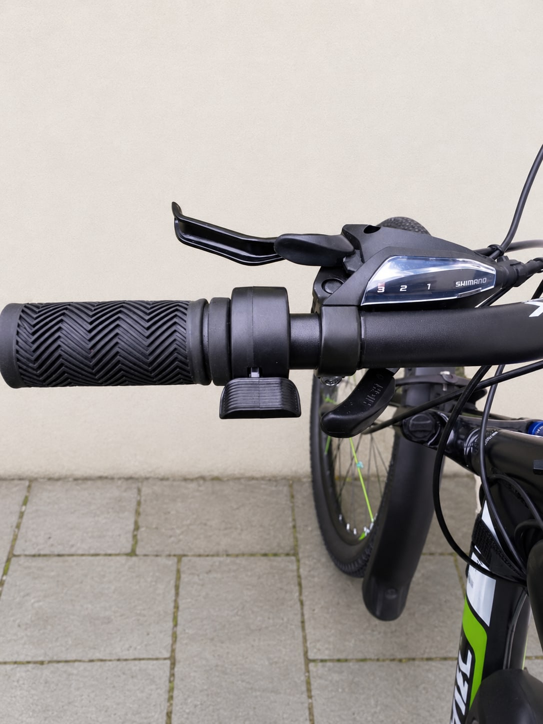Close-up of black Shimano shifter and grip on bicycle handlebars, non-drive side, against a light stone wall.