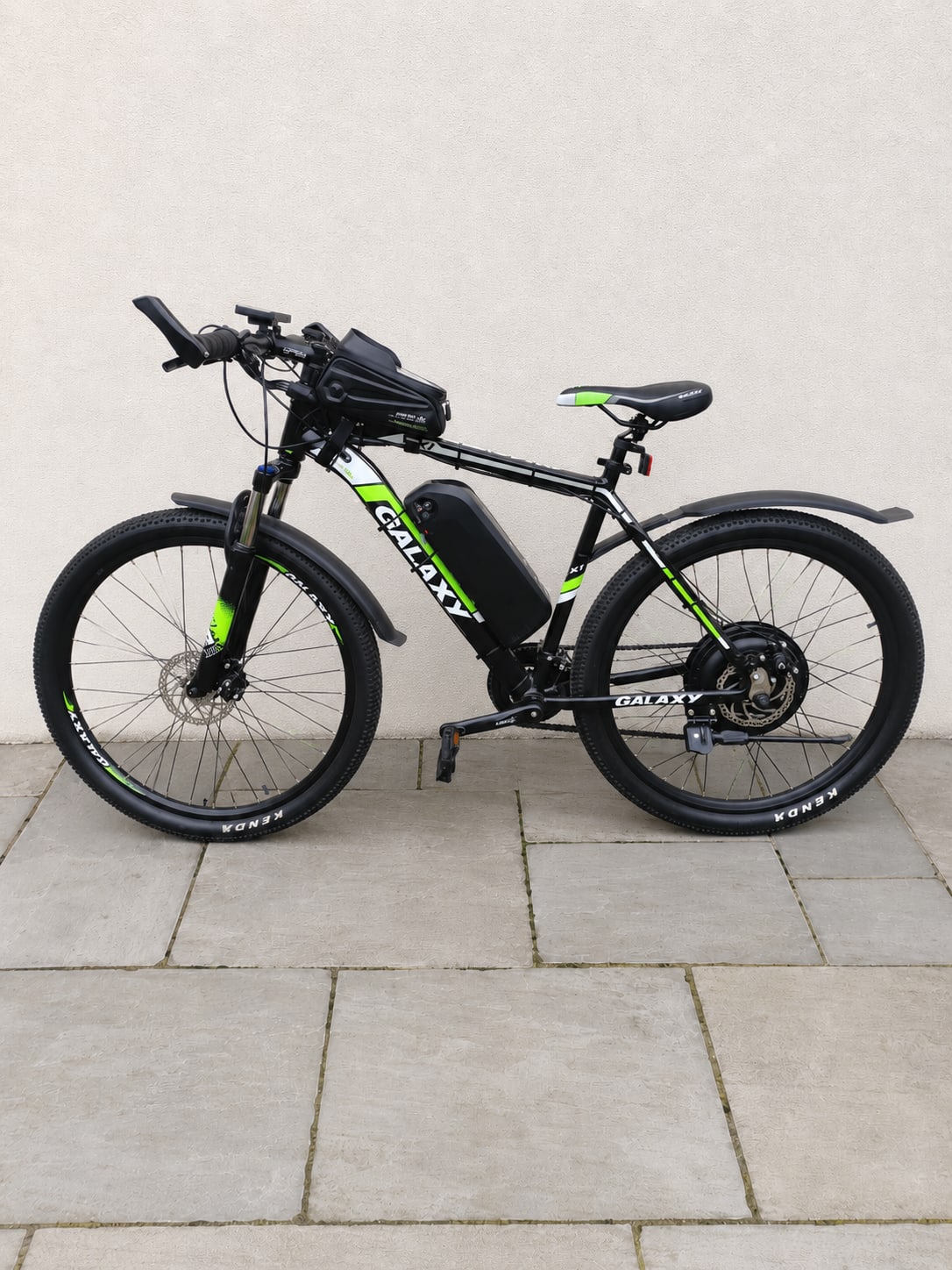 Black and neon-green electric mountain bike with front suspension and disc brakes, drive side, on paved ground against a white wall.