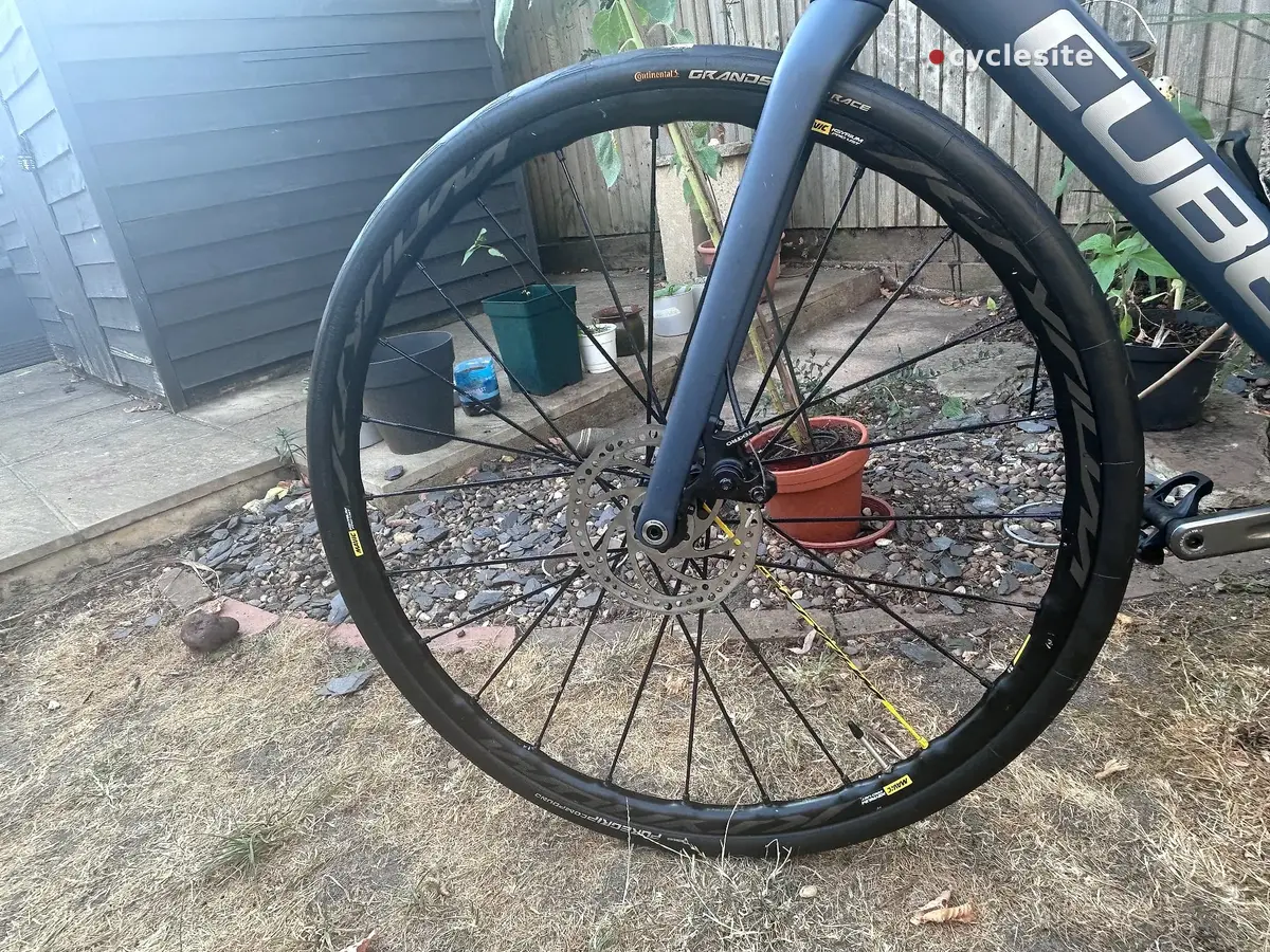 Front wheel of blue gravel bike with Continental Grand Prix tyres, close-up on drive side, outdoor garden setting with shed and gravel ground.