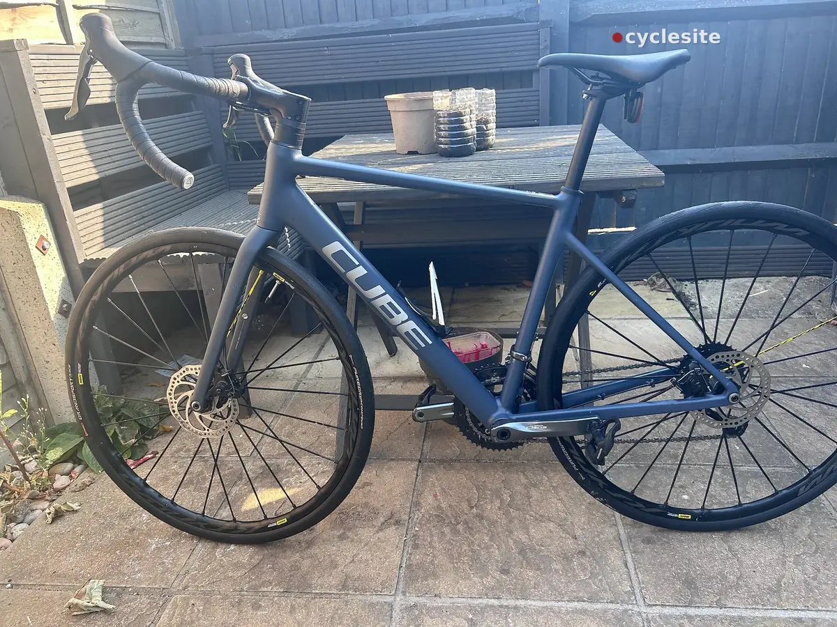 Blue Cube road bike with drop bars, parked on concrete outside a metal shed, drive side view, with red rear light visible.