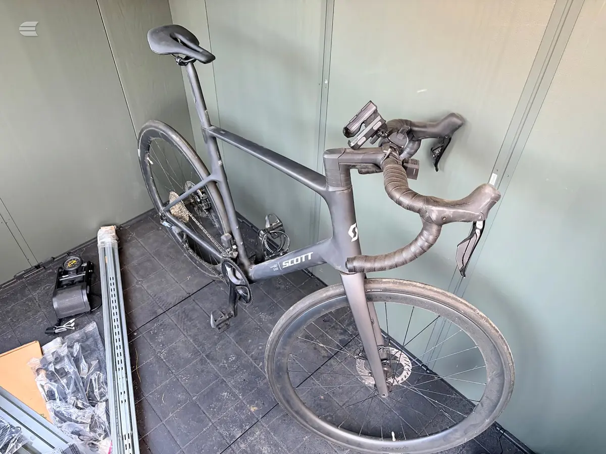 Scott gravel bike, grey, non-drive side, leaning against white wall in storage area with shelving visible.